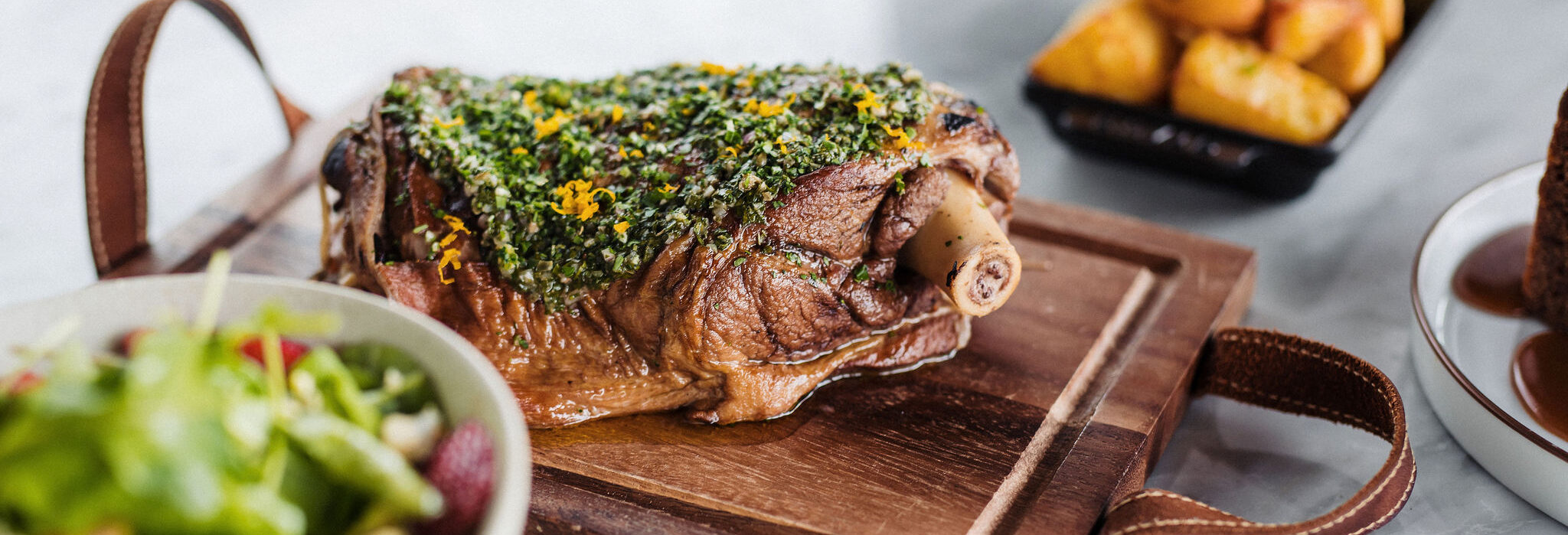 a roasted lamb shoulder sitting on a wooden cutting board with crispy roast potatoes in a black bowl in the background and a small green salad in a white bowl to the left side of the table.